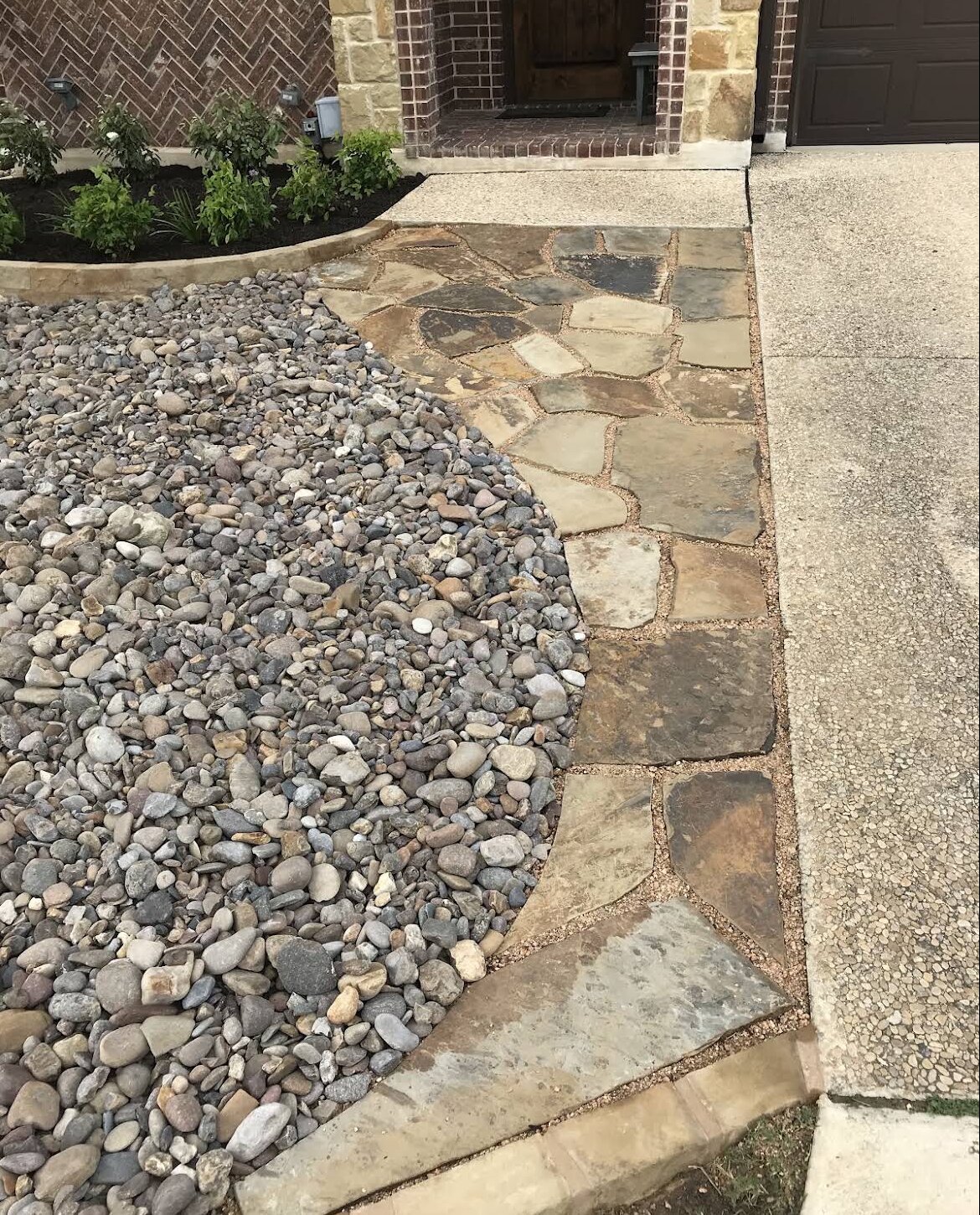 curved flagstone walkway between river rock and concrete driveway toward brick house entrance